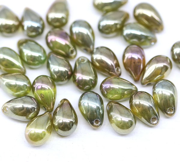 A group of small teardrop-shaped glossy beads with an iridescent olive green and gold finish, scattered on a plain white background.